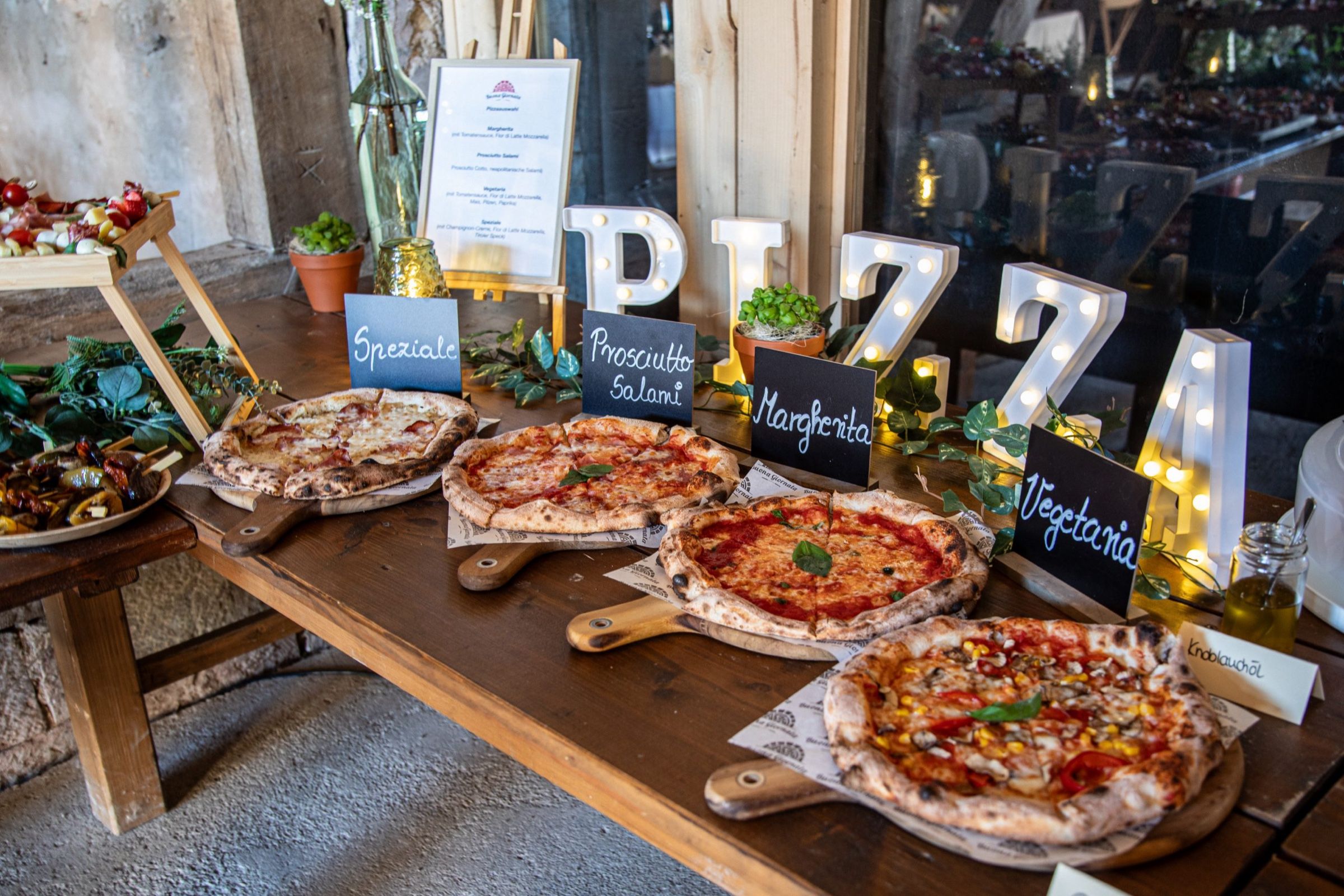 Vier Pizzen auf Holzbrettern mit Kreidetafeln, dahinter Leuchtbuchstaben „PIZZA“ und eine Pizzakarte