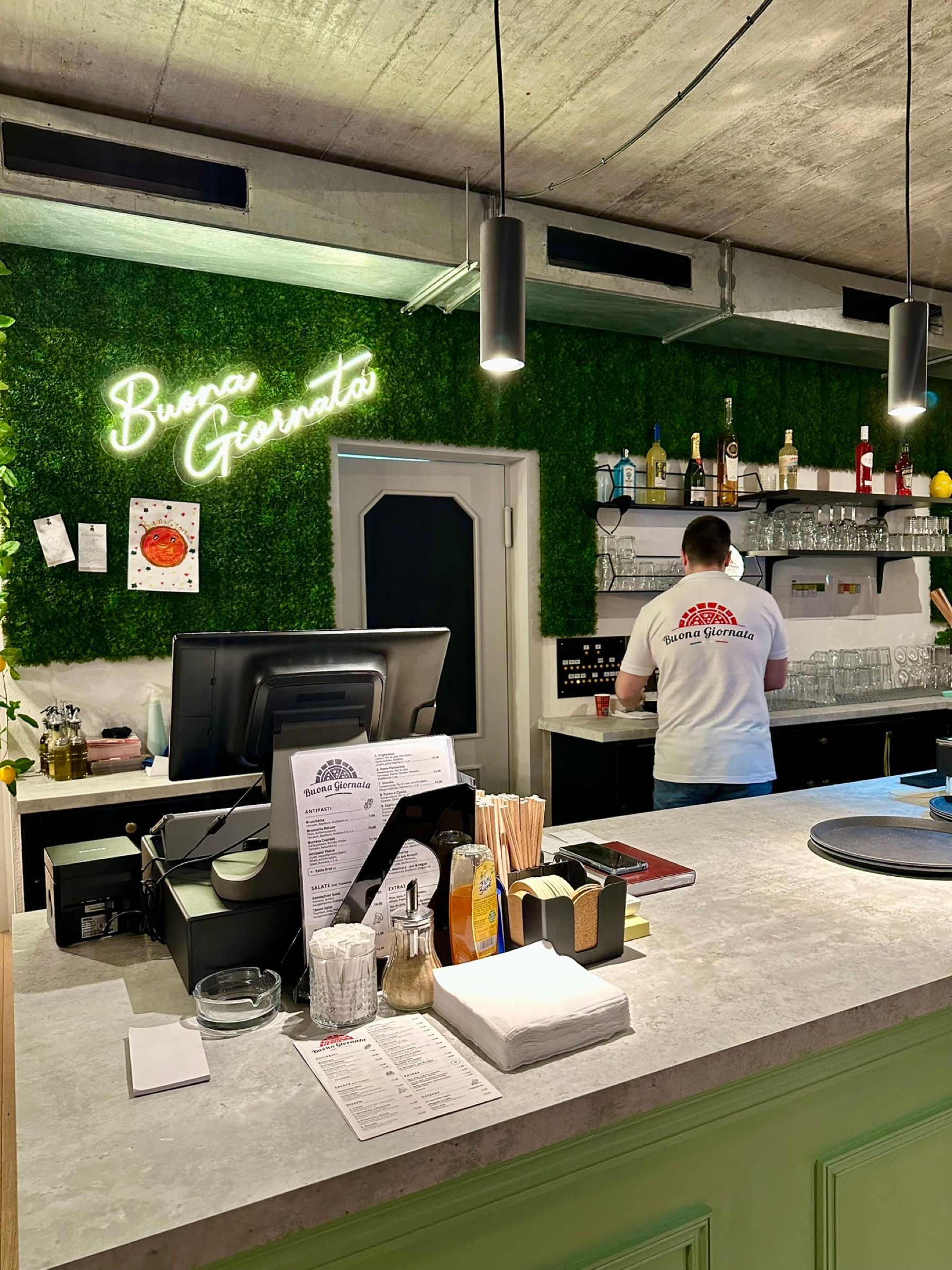 Theke der Pizzabar mit Neonschriftzug „Buona Giornata“ an einer grünen Pflanzenwand