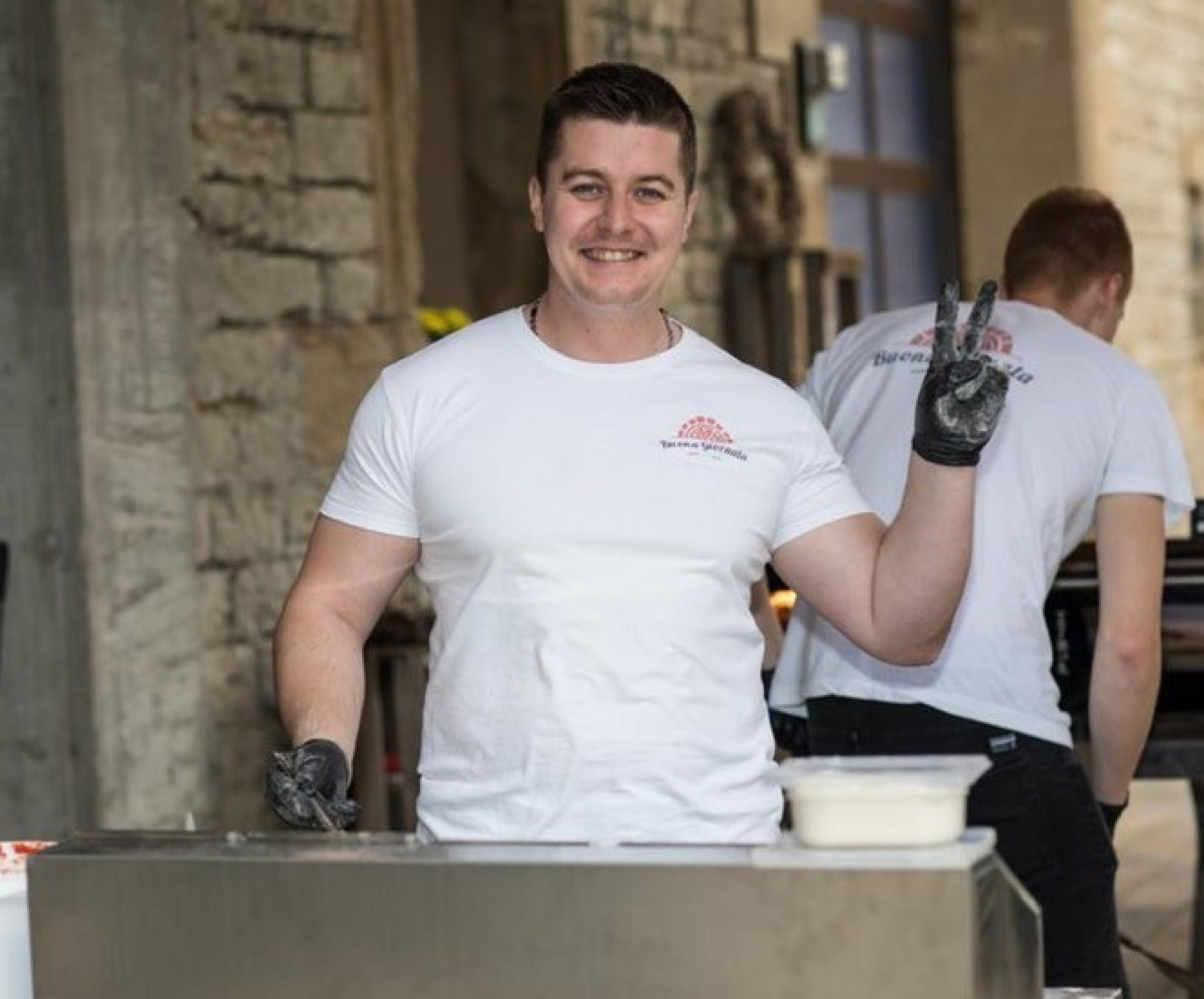 Lachender Mann im Buona-Giornata-T-Shirt mit schwarzen Handschuhen hinter einer Edelstahltheke, dahinter ein Kollege