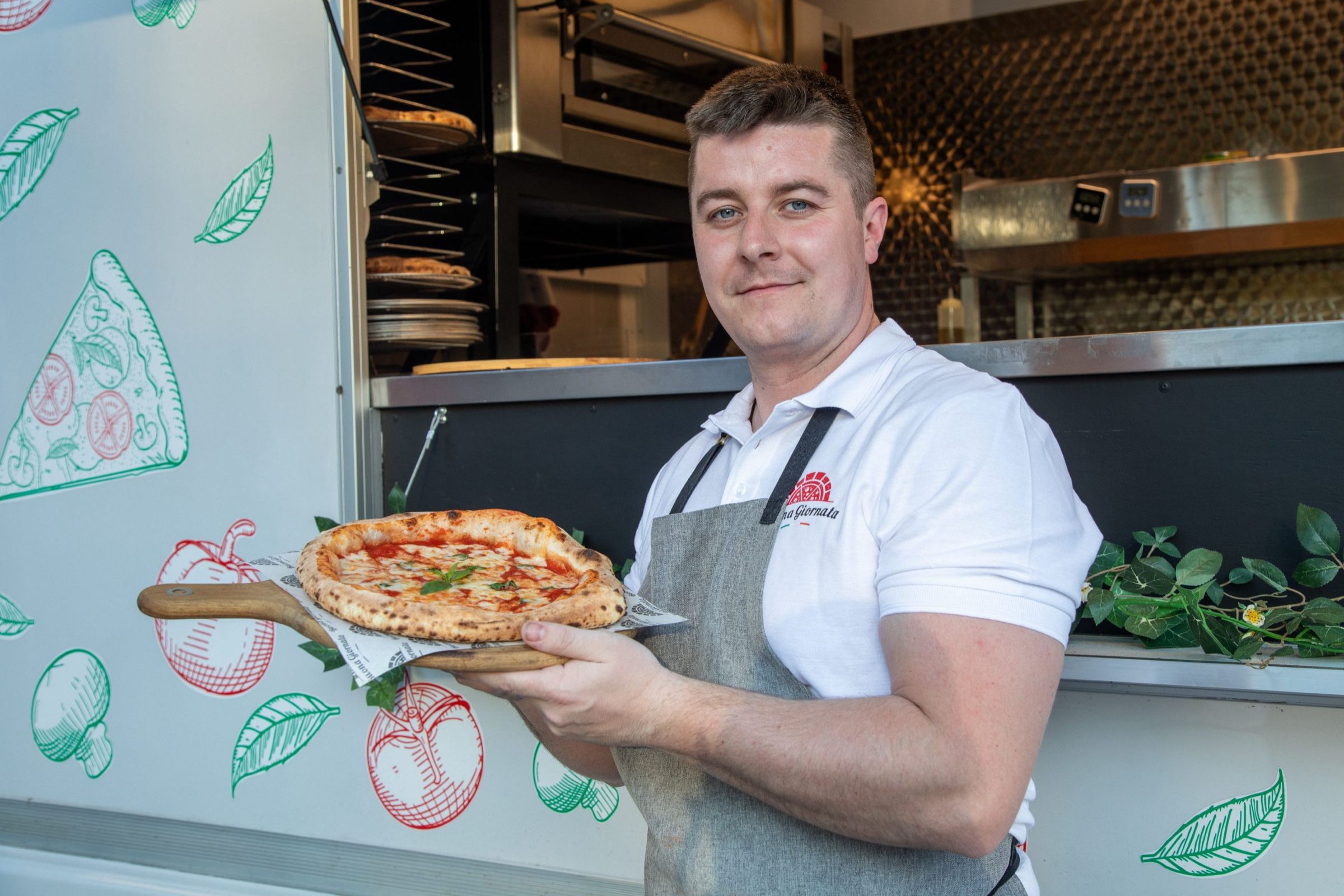 Mann mit Schürze hält eine Pizza auf einem Holzbrett vor der Ausgabe des Pizza Trucks
